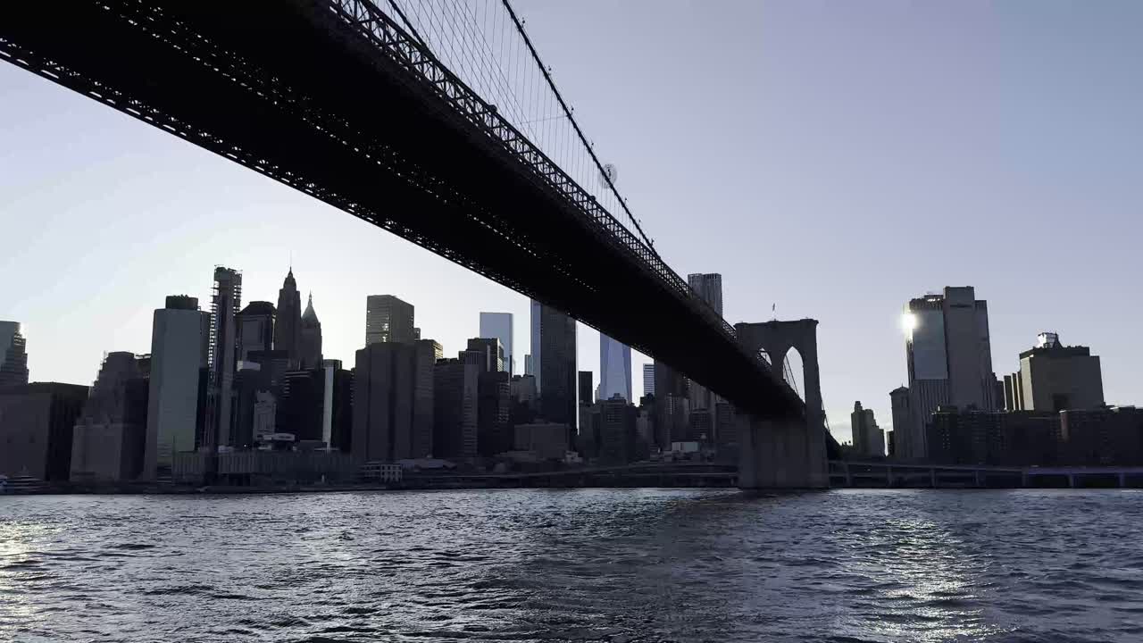 En fengslende utsikt fra under Brooklyn Bridge i skumringen, som fremhever silhuetten av Manhattans skyline mot skumringshimmelen og det glitrende vannet i East River.