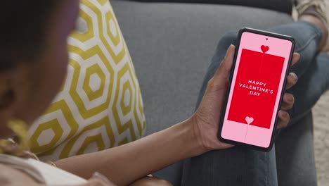 close up of woman sitting on sofa at home looking at happy valentine's day message on mobile phone