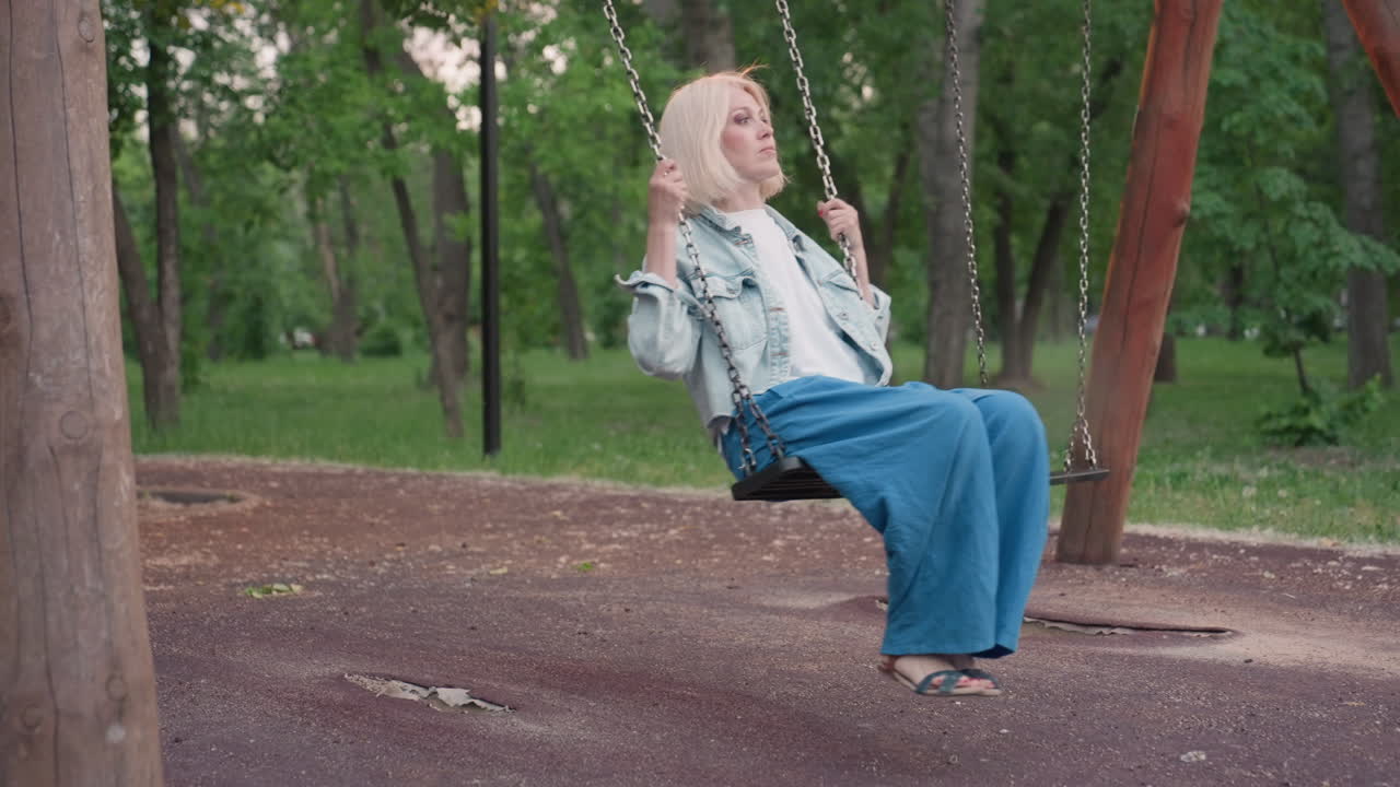 female parent seated on swing bench in quiet park dusk moment holding chains looking thoughtful surrounded by trees soft ambient light capturing contemplative solitude and movement under sky