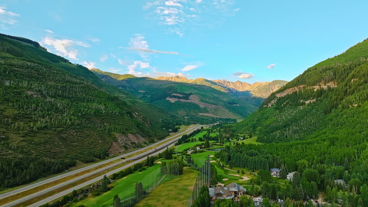 Drone tracking left tilt up along golf course of open Vail landscape with winding mountain road and sparse tree cover