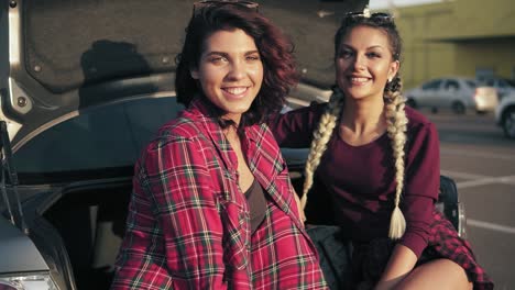 closeup shot of two young beautiful women sitting in a car trunk in the parking by the shopping mall during sunny day, smiling and looking in the camera