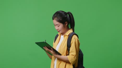 side view of asian woman student with a backpack smiling and taking note on the tablet while standing in the green screen background studio