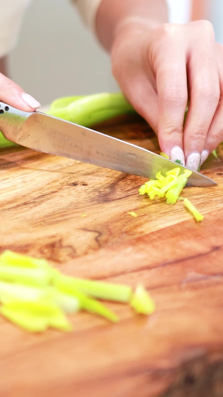 Primo piano di mani che tagliano abilmente il sedano su un tagliere di legno. L'illuminazione brillante evidenzia gli ingredienti freschi e la precisione del taglio.