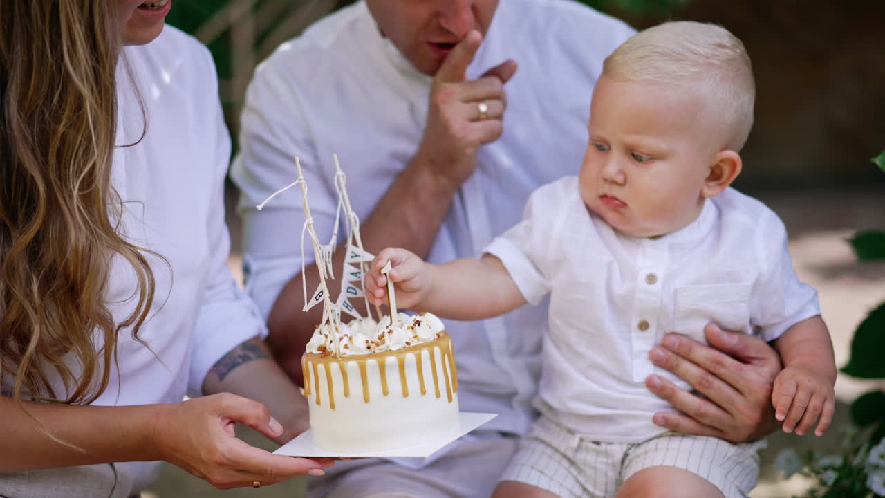 Parents sit holding a baby and a birthday cake in front of him. Serious blond baby boy takes number one decor from dessert. First anniversary of a child.