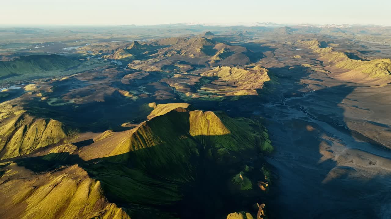 Vidsträckt drönarpanorama över Islands höglands vulkaniska öken med mossbeklädda toppar och svarta sanddalar, gyllene timmens ljus och långa skuggor som avslöjar en vidsträckt, robust terräng