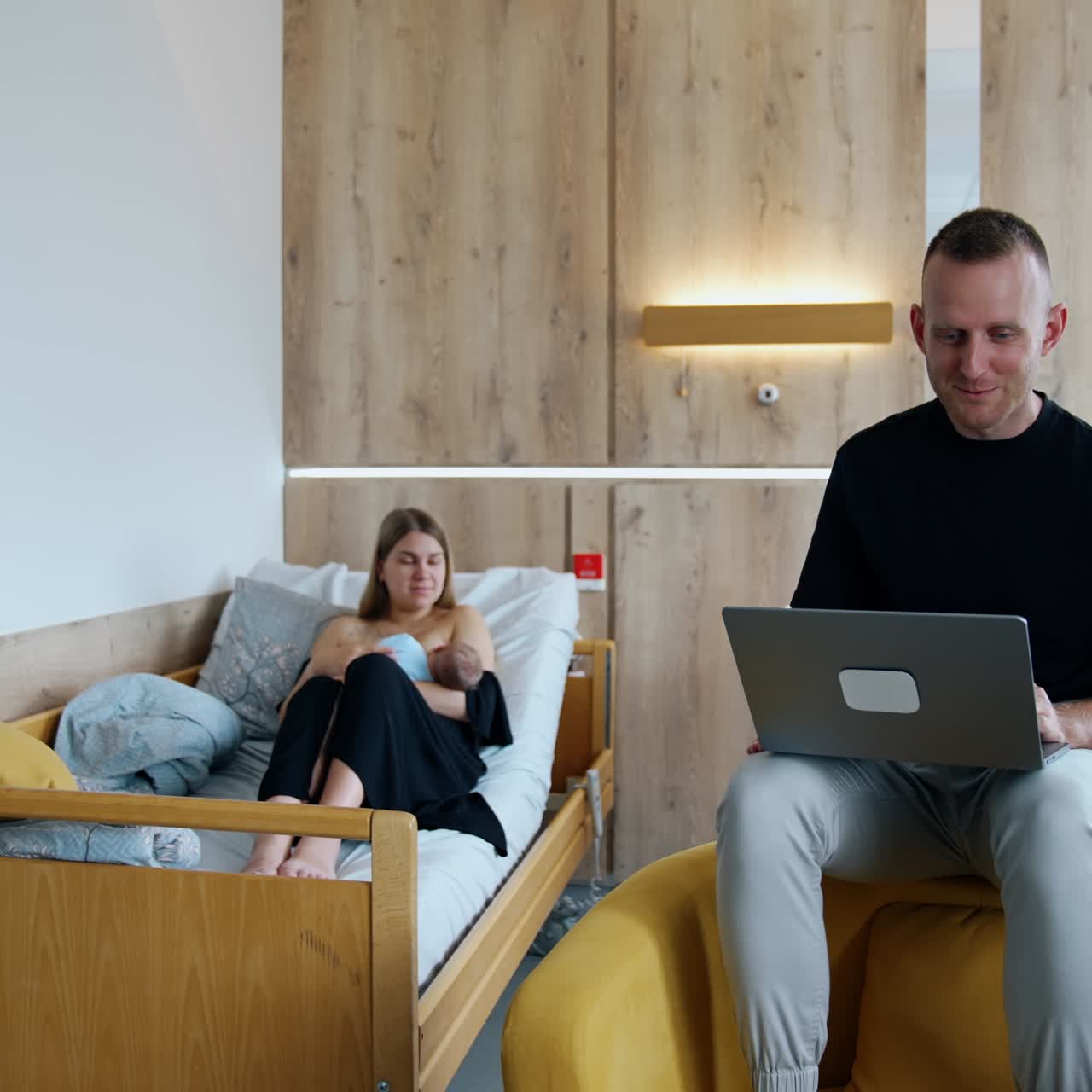 Married couple in the maternity hospital ward. Woman takes care of a baby and her husband works on laptop