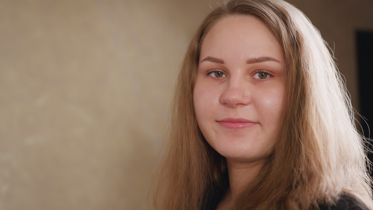 Zoom in portrait of student in black gown with front zipper, eyes closed, adjusting hair and posing with warm smile in soft indoor light, confident calm vibe against beige background