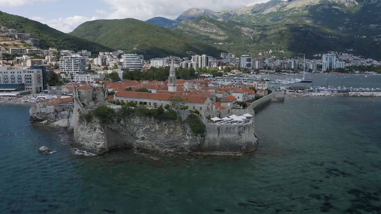Veduta aerea con drone della Cittadella di Budva e della Città Vecchia su una penisola rocciosa con mura in pietra ed edifici con tetti arancioni, circondata da chiare acque adriatiche e montagne