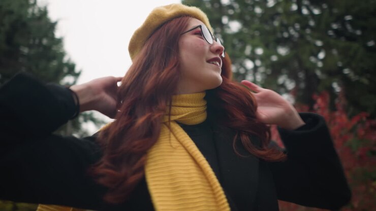 Splendida donna con capelli rossi ricci si volta gioiosamente, portando una mano alla testa, indossando un basco giallo, occhiali da sole e una sciarpa in un vibrante giardino dai colori autunnali.