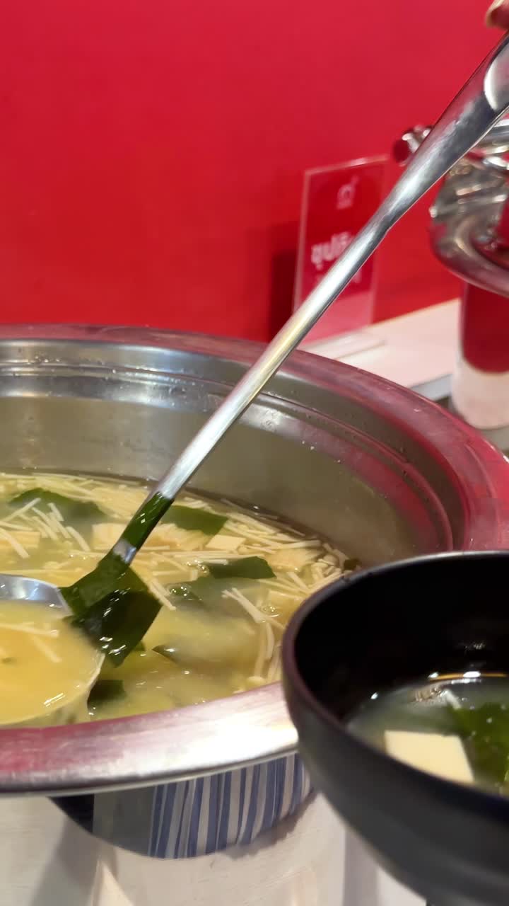 Una persona versa la zuppa di miso in una ciotola in un ristorante coreano BBQ a Bangkok. L'illuminazione brillante evidenzia gli ingredienti della zuppa.