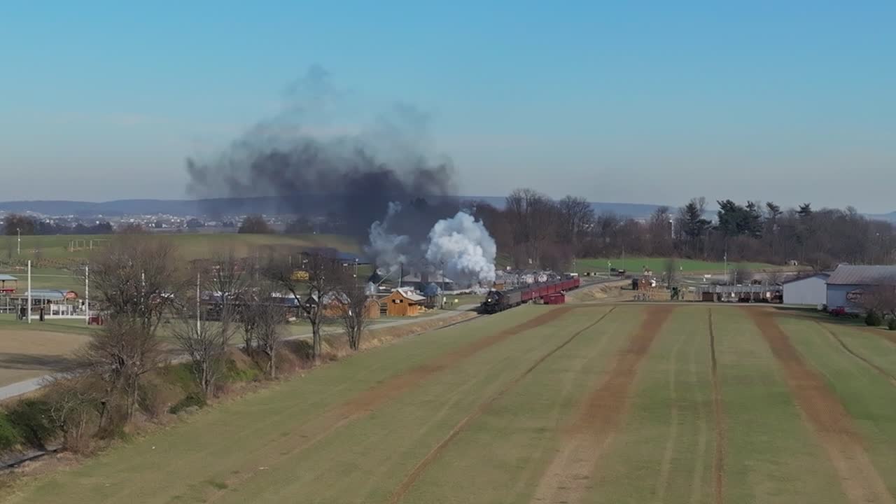 una vista aérea de un tren de pasajeros de vapor que se acerca, soplando humo, mientras viaja por el campo, en un soleado día de invierno