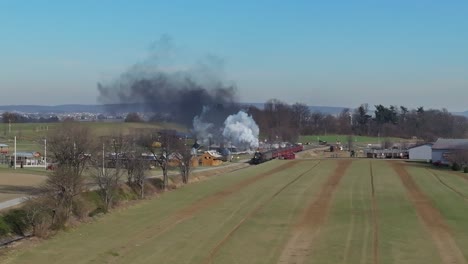 an aerial view of a steam passenger train approaching, blowing smoke, while traveling thru the countryside, on a sunny winter day