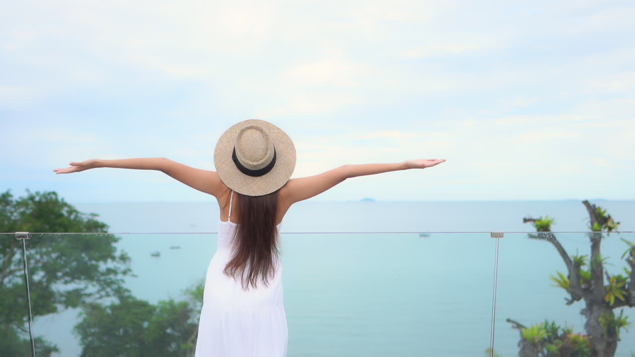 Back view of a female tourist walking towards balcony, looking at the ocean and rising her arms in the air enjoying her vacations
