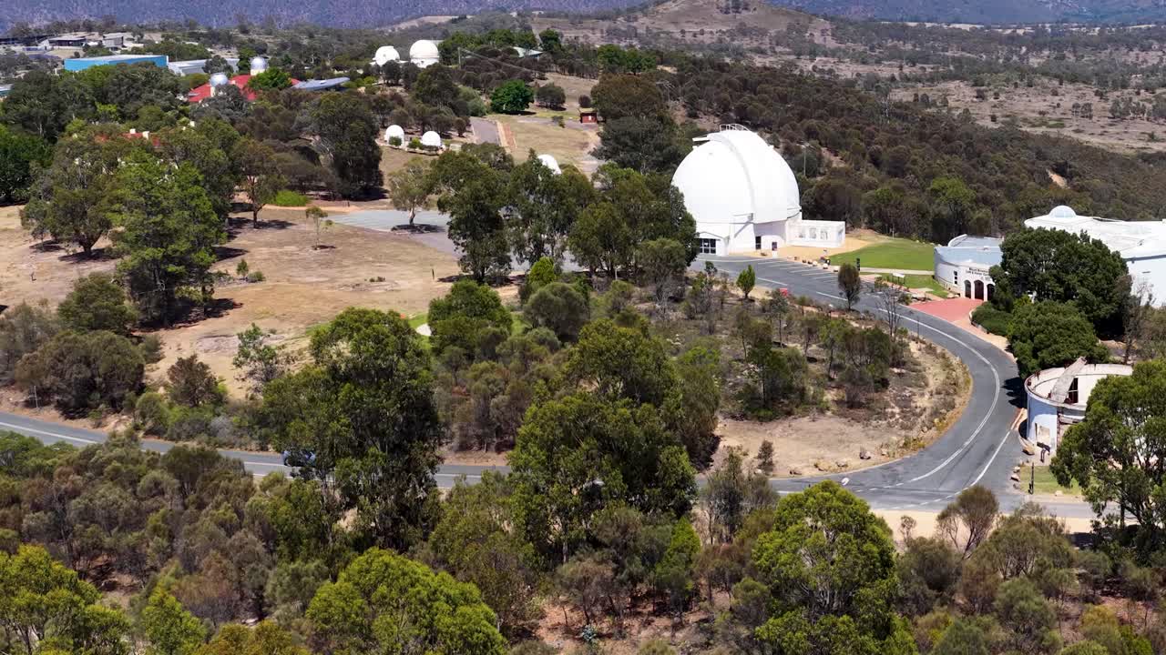 Bredt luftfoto med drone af hvide observatoriekupler på en skovklædt bakke i klart sollys
