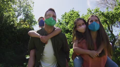 pareja caucásica con máscara facial llevando a un hijo y una hija en la espalda en el jardín