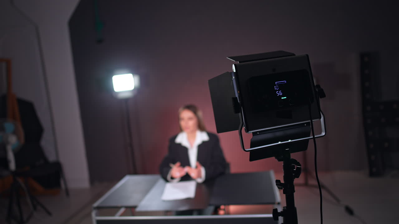 Lighting equipment back view at studio. Woman in black jacket and white shirt sits at blurred backdrop.