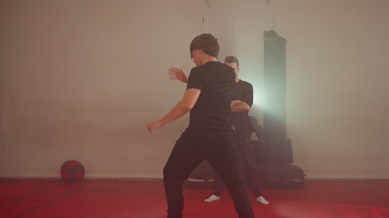 Karate practitioner performing dynamic movement during sparring training in gym, arms raised in motion on red mats, opponent standing in background, martial arts discipline with focus, under bright light