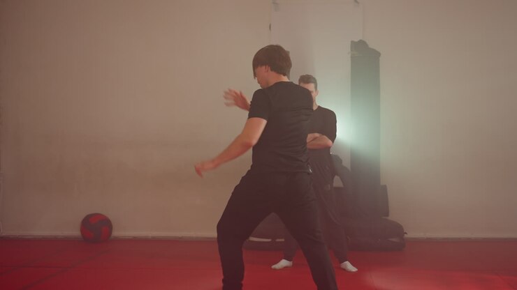 Praticante di karate che esegue un movimento dinamico durante un allenamento di sparring in palestra, braccia alzate in movimento su tappetini rossi, avversario sullo sfondo, disciplina di arti marziali con focus, sotto una luce intensa