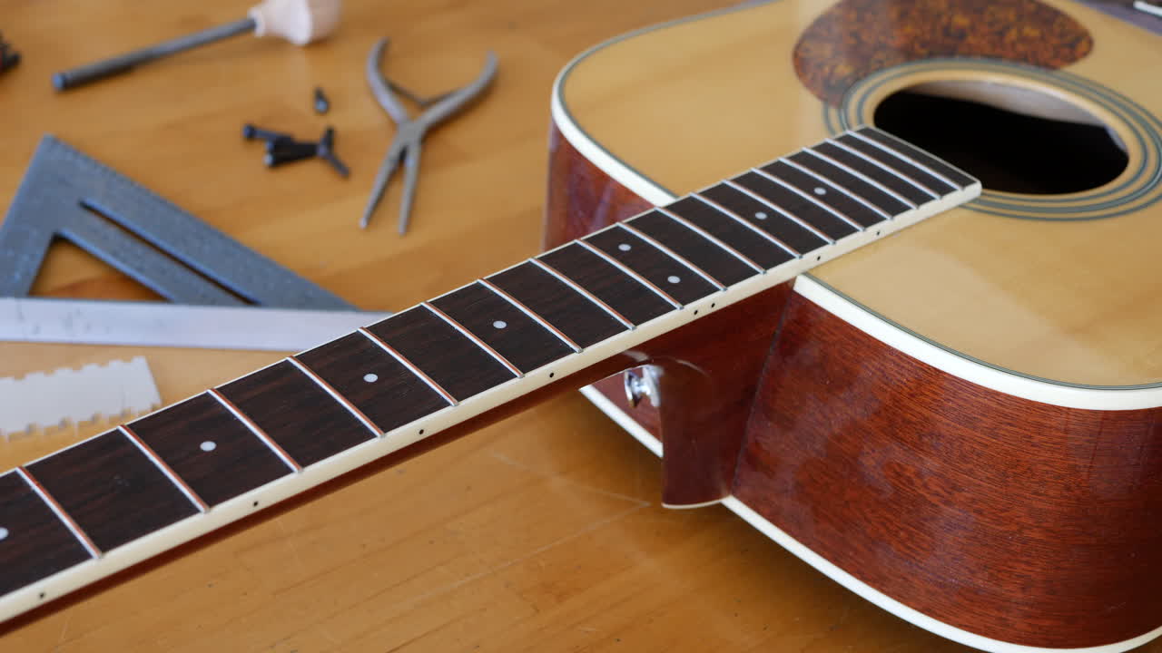 Close up hands of a luthier measuring, sanding and leveling the frets on an acoustic guitar neck on a wood workshop bench with lutherie tools