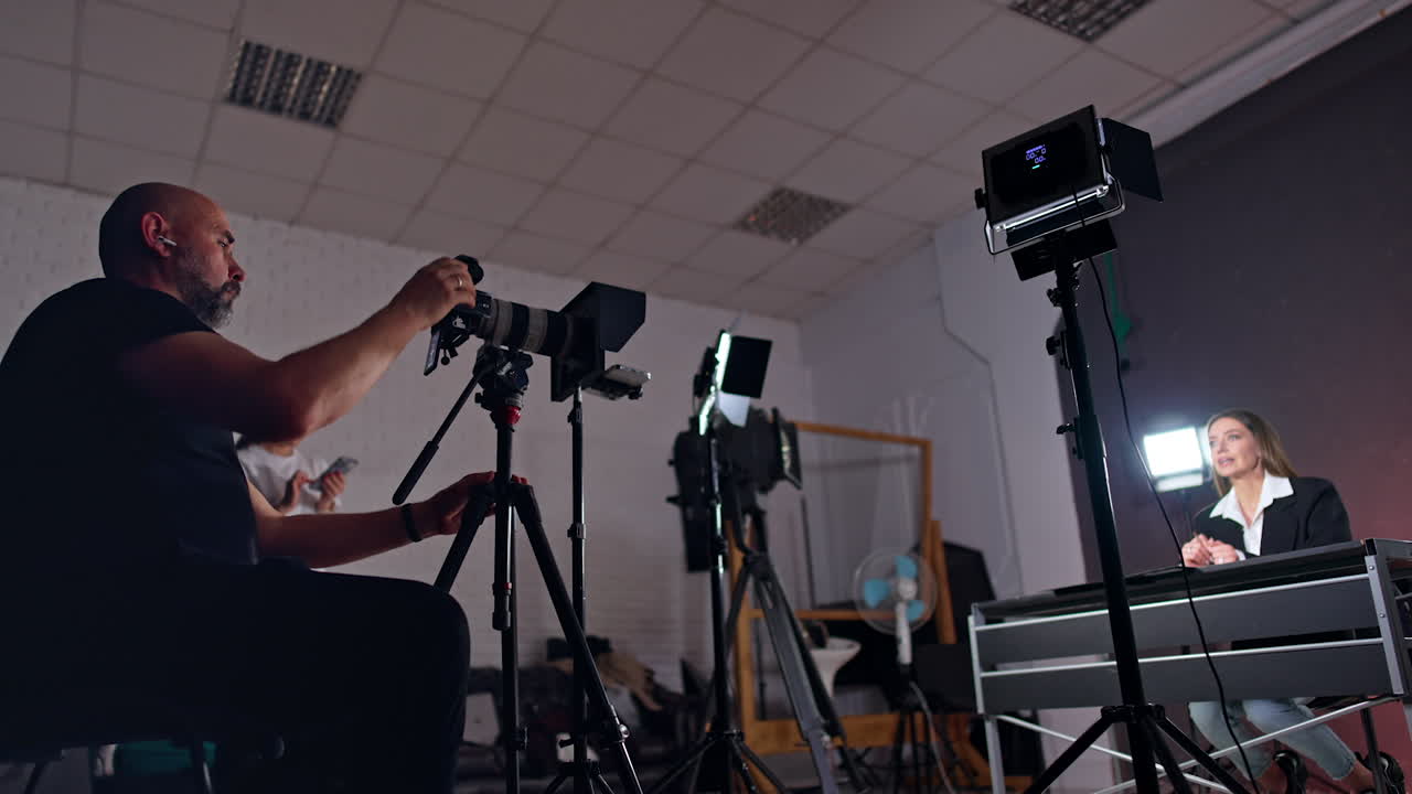 Bearded cameraman sits behind the camera recording a woman at desk. Low angle view. Girl with phone in hands stands at backdrop.