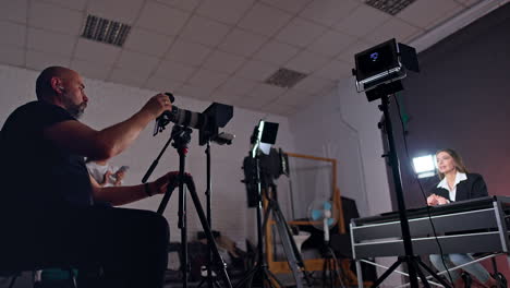 bearded cameraman sits behind the camera recording a woman at desk. low angle view. girl with phone in hands stands at backdrop.