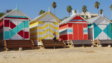 a women sitting on the stairs of a colourful beach box, sees someone she knows and waves to them