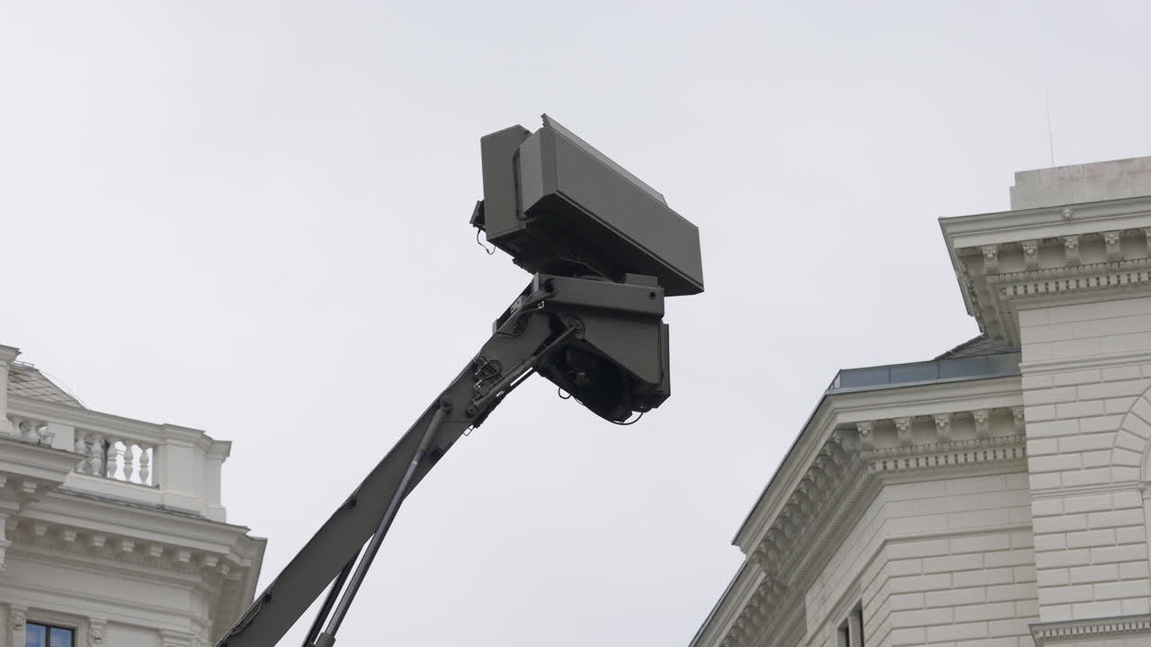 A large military radar or sensor array extends from an arm against an overcast sky, flanked by classical buildings