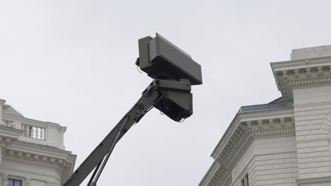 a large military radar or sensor array extends from an arm against an overcast sky, flanked by classical buildings