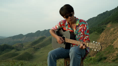 young male asian musician plays guitar in natural mountain landscape in vintage shirt jeans and sunglasses, sitting outdoors at nature