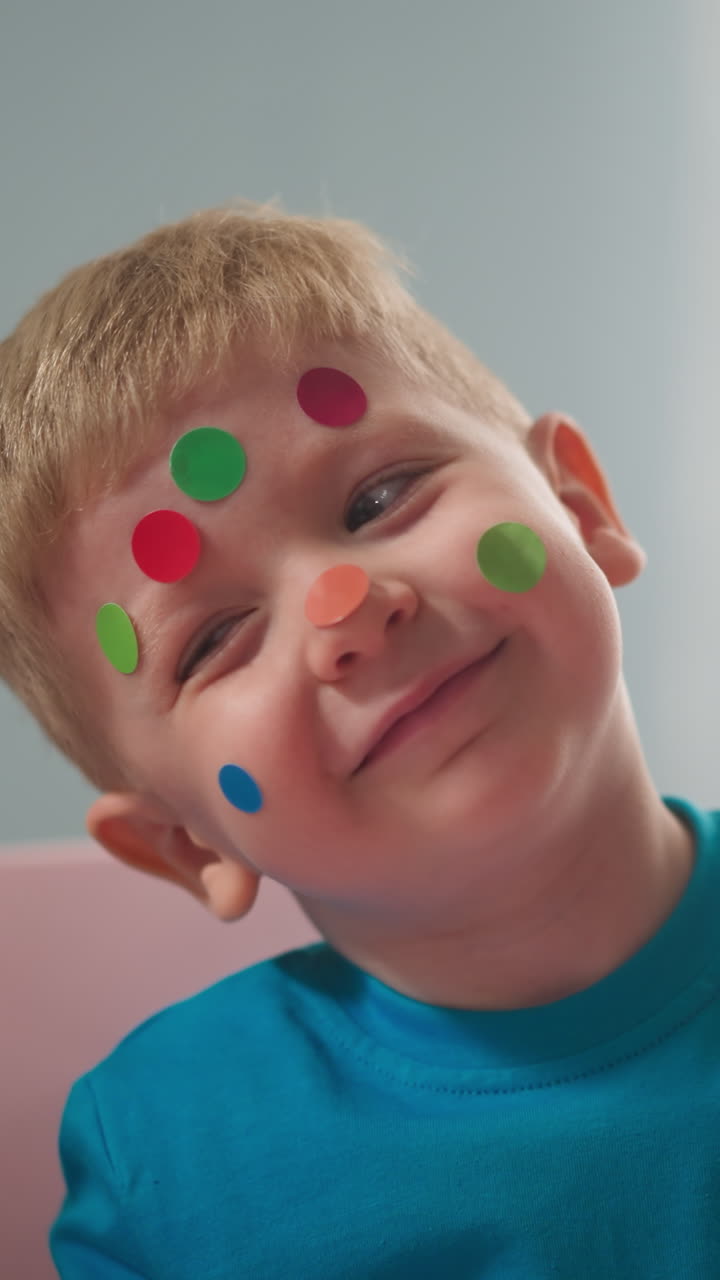 cheerful little child with multi-colored stickers in form of circle on his face laughs, looks at camera