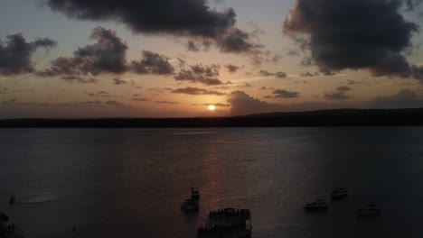 Dolly-out-aerial-shot-of-the-alligator-beach,-a-river-in-Cabedelo,-Paraiba,-Brazil,-near-the-coastal-capital-city-of-Joao-Pessoa,-with-tour-boats-watching-the-sunset-as-a-man-plays-the-saxophone-below