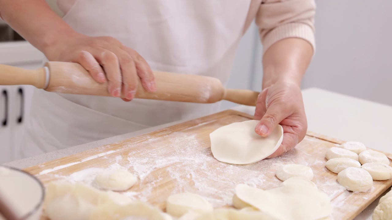 Unidentified hands of elderly woman chef wear apron preparing dough flattening it with rolling pin for bakery enjoy cookery in cozy kitchen. Women hands make dumplings from dough with meat on table