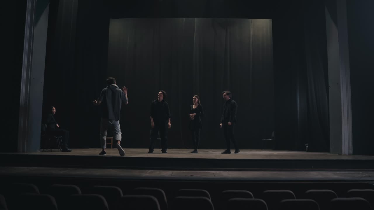 un director de escenario confiado con una camiseta gris sube al escenario para explicar a los actores cómo jugar y moverse correctamente en el escenario durante la preparación para una actuación en un teatro vacío con cortinas negras