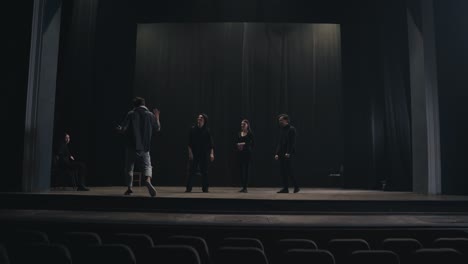 a confident stage director in a gray t-shirt comes on stage to explain to the actors how to play and move correctly on stage during preparation for a performance in an empty theater with black curtains