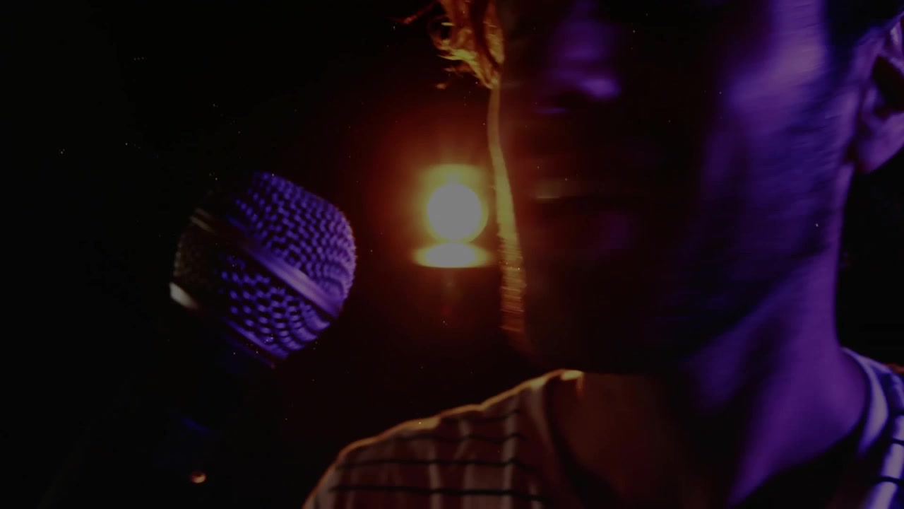 Cantante uomo che canta con illuminazione viola, fuochi d'artificio che scoppiano vicino al microfono mostrando l'energia della musica.
