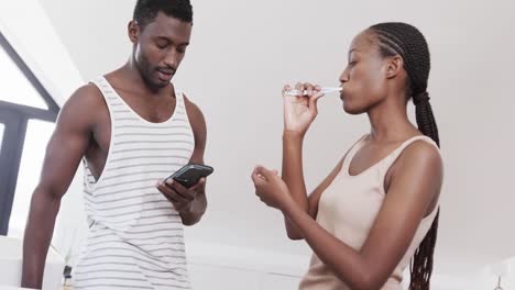 pareja afroamericana cepillándose los dientes, usando el teléfono inteligente y hablando en el baño, cámara lenta