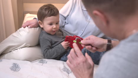 Baby,-Das-Auf-Dem-Schoß-Seiner-Mutter-Auf-Dem-Bett-Liegt,-Während-Es-Mit-Seinem-Vater-Spielt