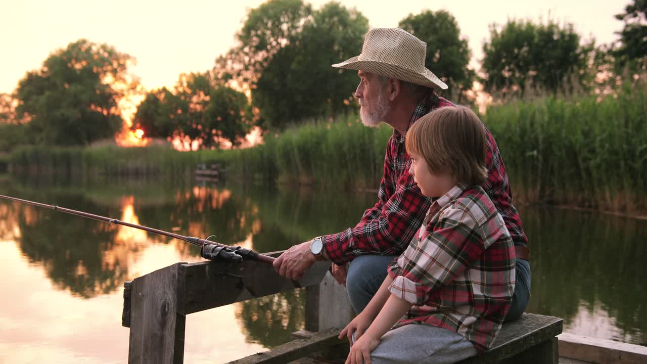 Close-up view of a grandfather and his grandson fishing and talking sitting on the lake pier on a summer day at sunset