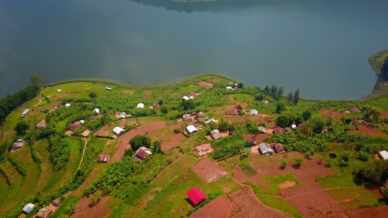 Aerial orbit reveals a patchwork of terraced farmland, scattered tin-roofed homes, and cultivated green plots descending toward the shore of Lake Bunyonyi in southwestern Uganda