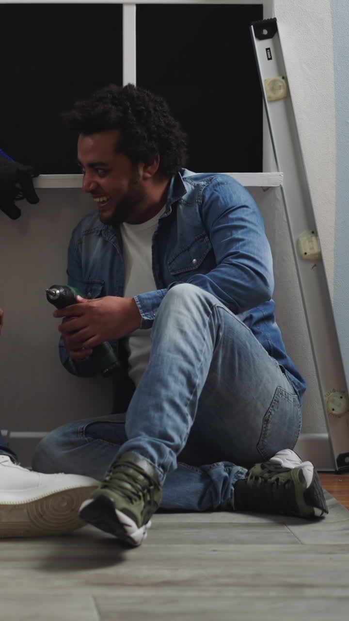 Joyful African-American man shows wireless screwdriver with torch to friend at talk sitting near window. Workers check electric tool for repair in evening