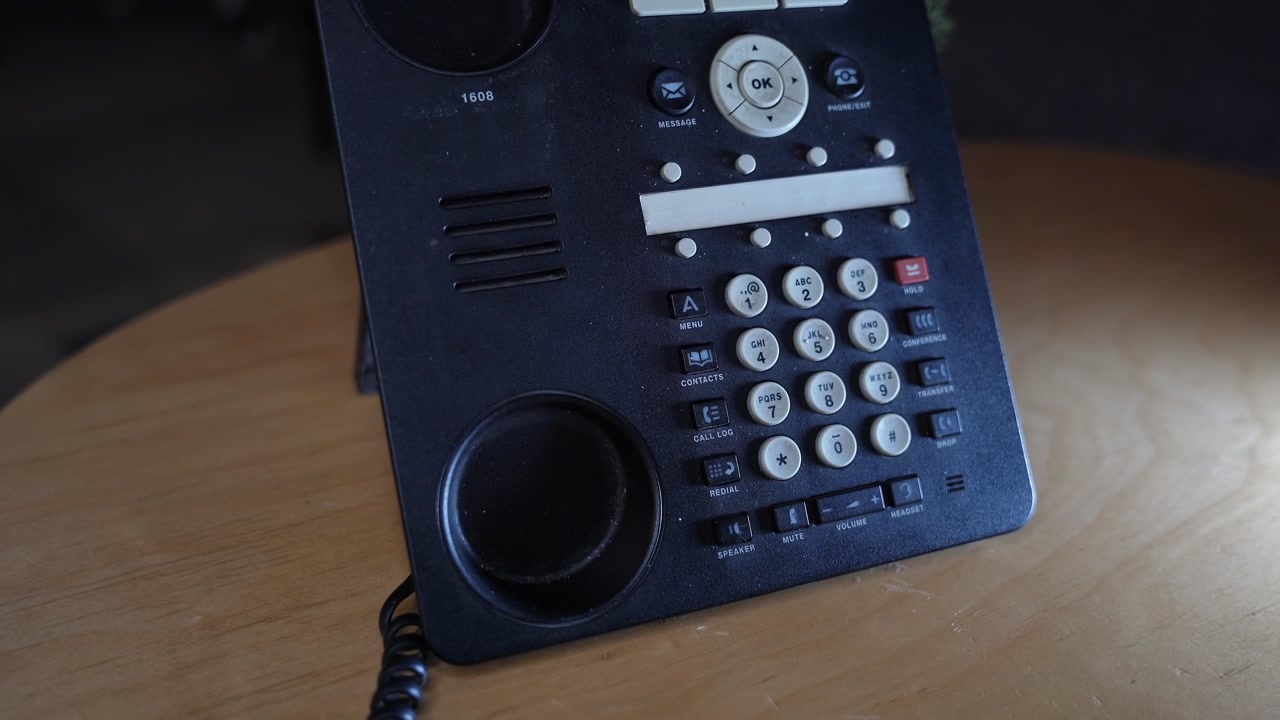 A close-up shot of a person's hand picking up the receiver and dialing a number on a black landline telephone. The device sits on a wooden desk in a dimly lit indoor office setting.
