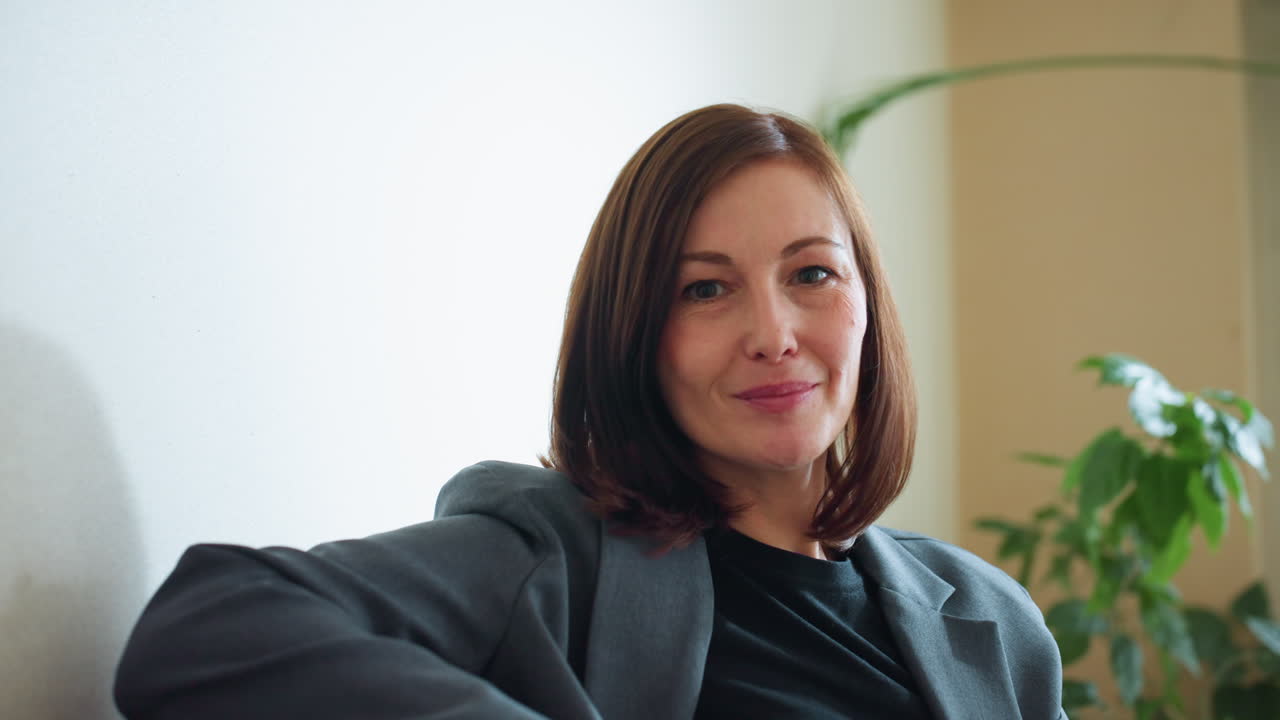 Professionista in blazer grigio seduta comodamente su un divano in un ufficio luminoso con piante da interno, che sorride dolcemente e trasuda calma e sicurezza in un ambiente di lavoro rilassato con luce naturale