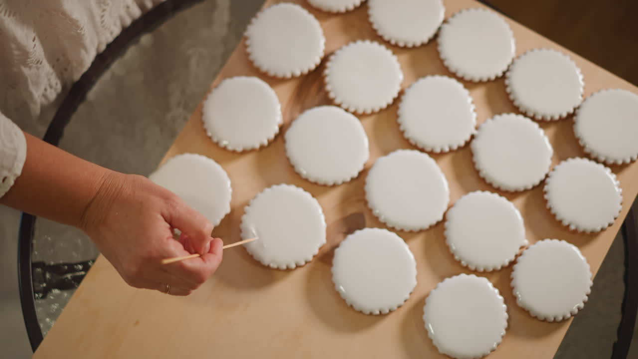 Caucasian hands decorating iced cookies on wooden board, closeup of precise toolwork smoothing fondant tops across rows of round treats, neutral warm light, artisanal baking routine and careful