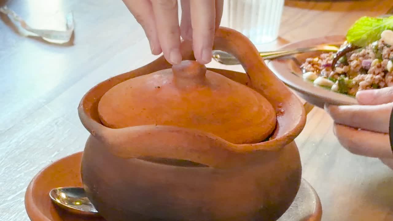 Hand lifts clay pot lid, unveiling steaming Thai soup in bright, casual dining setting