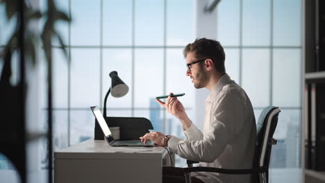 Young-Guy-Talking-On-Mobile-Phone-And-Taking-Notes-Using-Voice-Assistant-Writing-In-Notebook.-Smiling-Man-Sitting-At-Desk-With-Laptop-At-Home-Office-Wearing-Earbuds