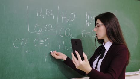 escolarización en línea. la maestra está dando una lección de química en línea para los estudiantes, remotamente, usando un teléfono inteligente. está escribiendo fórmulas con tiza en la pizarra, en la clase escolar. enseñanza remota. transmisión de video en vivo desde la clase