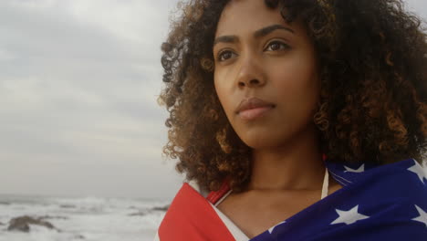 vista frontal de una mujer afroamericana envuelta en una bandera estadounidense de pie en la playa 4k