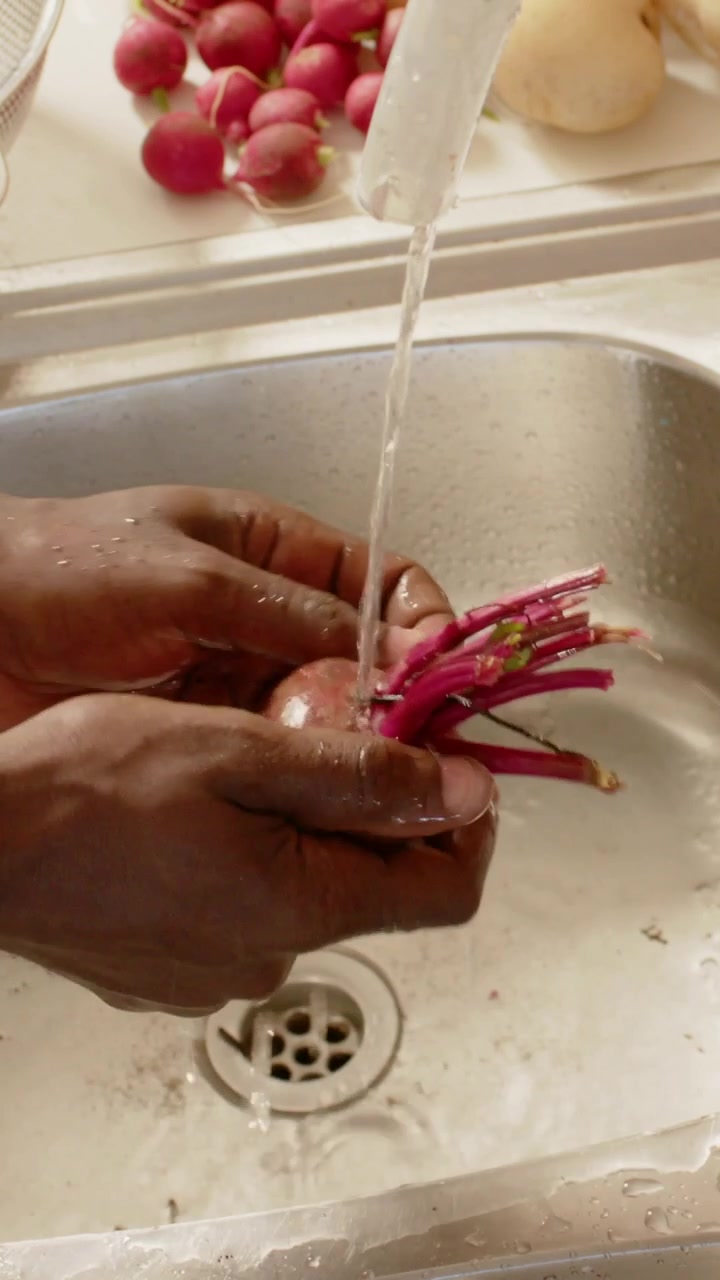 Vertical video: Rinsing hands trimming radish under running faucet at kitchen sink, removing dirt