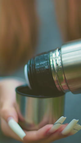 close-up of someone with well polished nails pressing thermos cover to pour water into lid, against blurred greenery background, highlighting careful handling of drink outdoors