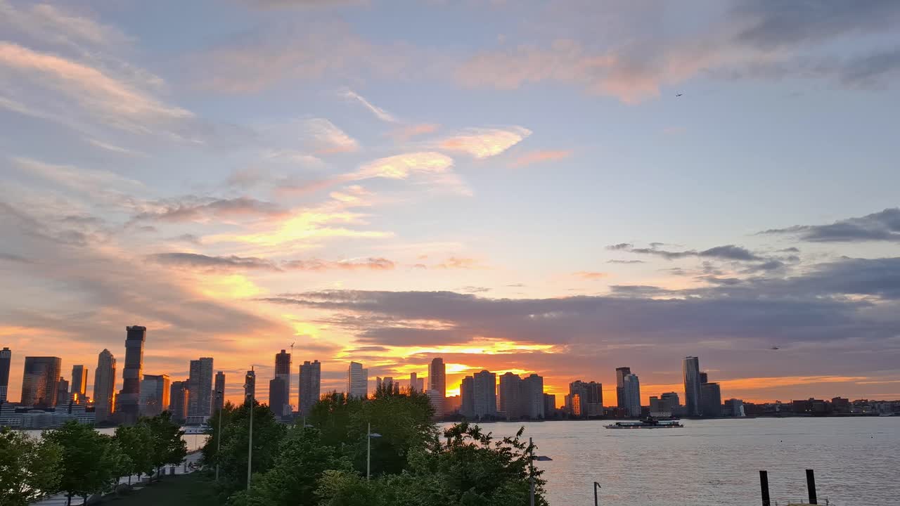 A wide view of the city skyline with tall buildings reflecting sunset light across the river, with trees and a waterfront area in New York, United States